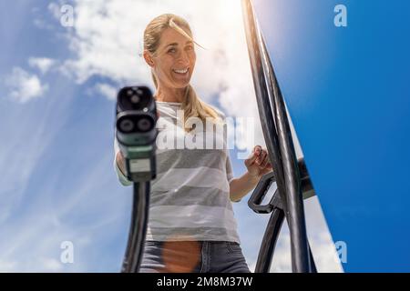 Donna felice che tiene un connettore a spina di ricarica DC CCS2 EV su Hypercharger o Supercharger stazione di ricarica per ricaricare la sua auto. Foto Stock