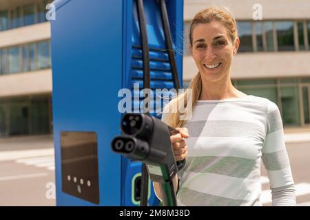 Bella donna che tiene un connettore di carica DC CCS2 EV su un Hypercharger o Supercharger per ricaricare la sua auto. Foto Stock