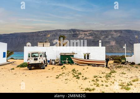 Paesaggio urbano di Caleta del Sebo, strade sabbiose, auto 4x4, barche e case bianche con l'isola vulcanica di Lanzarote sullo sfondo, la Graciosa, Isole Canarie Foto Stock