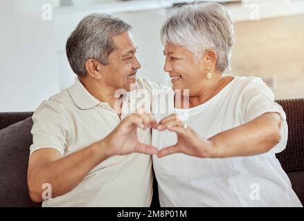 Sorridente coppia anziana di gara mista che fa forma al cuore, segno, simbolo con gesto della mano a casa. Felice marito ispanico e moglie seduti insieme Foto Stock