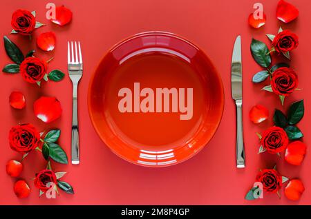 Cucina di San Valentino con piatto rosso, forchetta e coltello messi su tavolo rosso con rose, petali e foglie. Foto Stock
