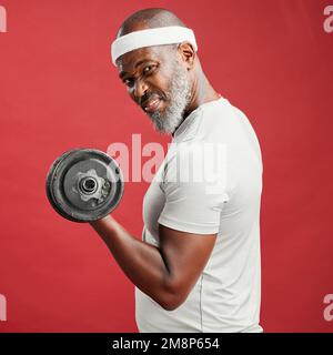 Uomo africano-americano maturo sicuro che si alza da solo contro uno sfondo rosso in uno studio e posa con manubri. Un uomo nero concentrato che si sente sportivo Foto Stock