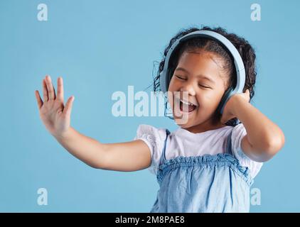 Adorabile bambina ispanica in piedi con gli occhi chiusi e felice di ascoltare musica con cuffie wireless, ballare e cantare Foto Stock