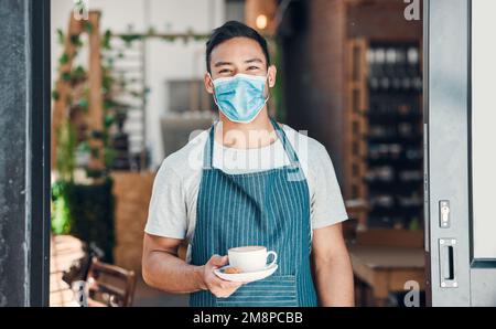 Ritratto di un giovane cameriere ispanico che indossa una maschera facciale e serve una tazza di caffè mentre lavora in un bar. Un barista cordiale che segue la sicurezza Foto Stock