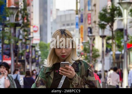 Una donna giapponese, con capelli biondi tinti, che usa un iPhone per strada a Ikebukuro, Tokyo, Giappone. Foto Stock