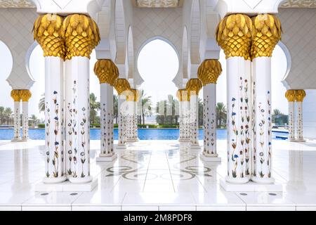 Un colonnato di marmo bianco con capitelli dorati che conduce alla piscina esterna con palme sullo sfondo della moschea Sheik Zayed Foto Stock