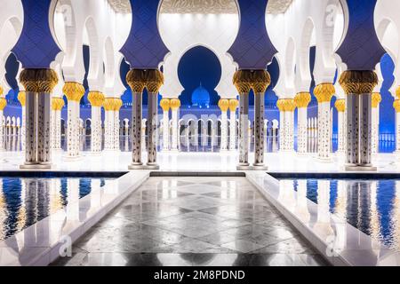 foto notturna simmetrica del colonnato della moschea dello sceicco Zhayed, con una passerella in marmo circondata dall'acqua Foto Stock