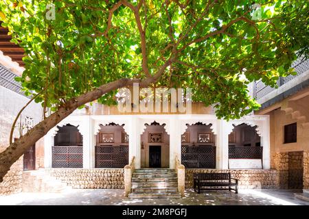 Antica casa tradizionale nella vecchia Dubai, con un portico fiancheggiato da colonne di marmo che forniscono ombra e un albero di quercia in primo piano Foto Stock