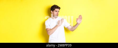 Uomo che mostra abilità kung-fu, arti marziali movimento ninja, in piedi in bianco t-shirt pronto a combattere, in piedi su sfondo giallo Foto Stock