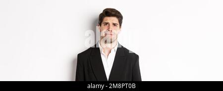 Primo piano di un uomo confuso e bello in tuta, grimacing e aspetto perplesso, vestendo abiti alla moda, in piedi su sfondo bianco Foto Stock