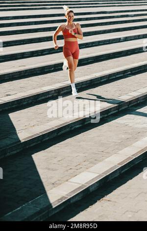 Runner femmina che si allenano giù per le scale all'aperto. Donna sportiva che fa un esercizio cardio al mattino. Donna caucasica felice che si sta allenando in sportswea Foto Stock