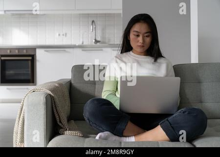 Una donna coreana professionista concentrata siede sul divano con un computer portatile e scrive testo in remoto Foto Stock