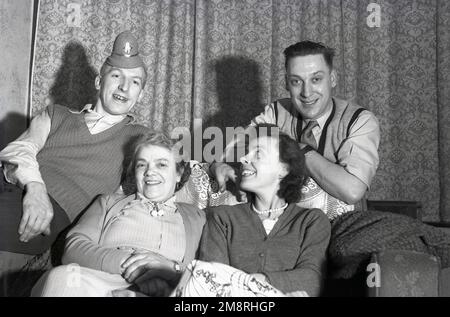 1950s, storico, foto di famiglia, due madri sedute su un divano con i loro figli, ridendo, come uno dei giovani si sta imbrogliando, indossando sulla sua testa un cappello da partito di un piccolo poliziotto, Inghilterra, Regno Unito. Foto Stock