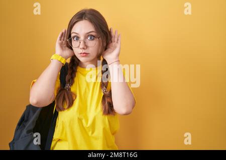 Giovane donna caucasica che indossa lo zaino da studente su sfondo giallo cercando di sentire entrambe le mani sul gesto auricolare, curioso di pettegolezzi. problema uditivo, d Foto Stock