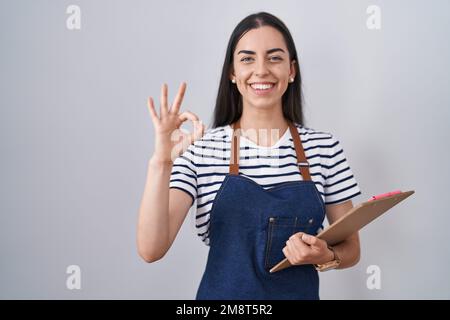 Giovane donna bruna indossando cameriera professionale grembiule e appunti facendo segno ok con le dita, sorridendo amichevole gesturing eccellente simbolo Foto Stock