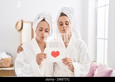 Donna e figlia di mezza età che indossa accappatoio che tiene carta del cuore fare pesce faccia con bocca e occhi squinting, pazzo e comico. Foto Stock