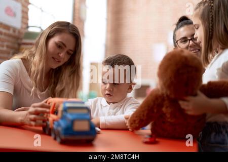 Insegnanti con ragazzo e ragazza che giocano con auto giocattolo e bambola seduti sul tavolo all'asilo Foto Stock