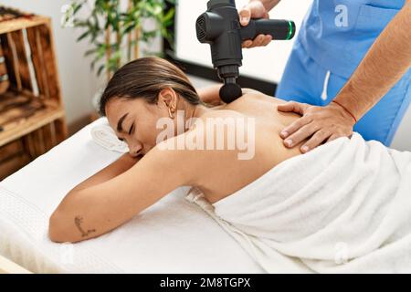 Uomo latino e donna che indossano l'uniforme di fisioterapia massaggiante indietro usando la pistola a percussione al centro di bellezza Foto Stock