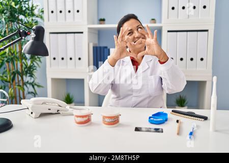 Donna dentista matura ispanica che lavora in clinica sorridendo innamorato mostrando il simbolo del cuore e la forma con le mani. concetto romantico. Foto Stock