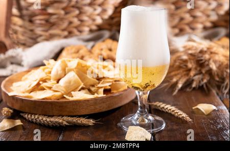 Bicchiere di birra artigianale leggera su un tavolo di legno. La birra viene versata in un bicchiere con schiuma e bolle. per i social media. consistenza della birra. Su un piatto ci sono patatine tradizionali in Italia in Sardegna Foto Stock