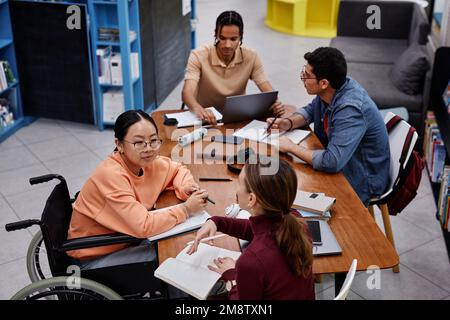 Vista ad alto angolo in diversi gruppi di studenti al tavolo in biblioteca, ragazza adolescente con disabilità che parla con un amico Foto Stock