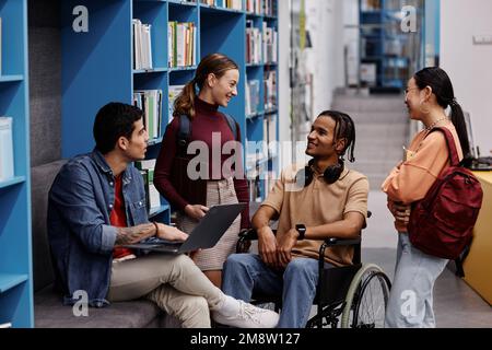 Gruppo vario di studenti con giovane in sedia a rotelle chattando allegramente in biblioteca universitaria, concetto di inclusività Foto Stock