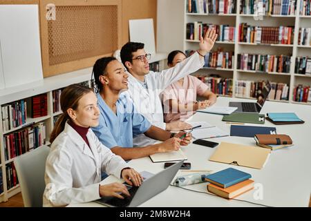 Gruppo di giovani studenti di medicinali che si siedono in fila e alzano le mani durante una lezione o un seminario Foto Stock