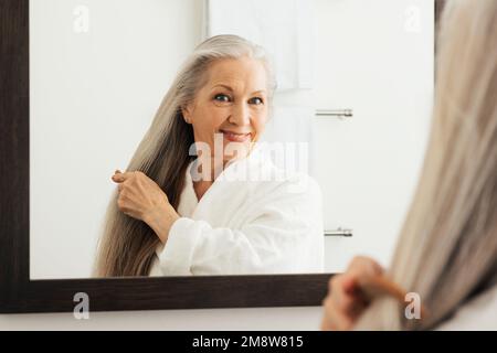 Donna matura che pettina i suoi lunghi capelli grigi davanti a uno specchio Foto Stock