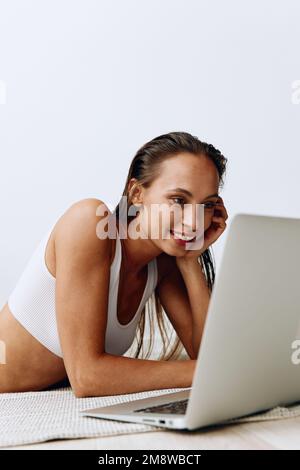 Una donna si trova sul pavimento a casa e sorride con i denti mentre guarda un notebook mentre chiacchiera online e guarda programmi TV durante la giornata Foto Stock