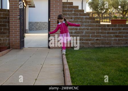 Immagine di una adorabile bambina che torna dalla scuola giocando a piedi su un marciapiede mentre si bilancia. Foto Stock