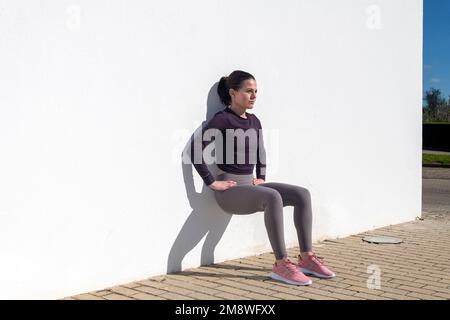 Donna sportiva che fa squat contro una parete bianca al sole. Foto Stock