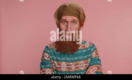 Divertente gioioso uomo hippie testa rossa sincero in camicia modello facendo divertenti espressioni facciali sciocco e grimacing, ingannando intorno mostrando la lingua. Ragazzo giovane e bello hipster isolato su sfondo rosa Foto Stock