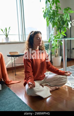 Giovane donna consapevole che indossa cuffie che fanno meditazione a casa. Verticale Foto Stock