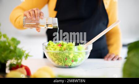 Giovane afroamericana in grembiule che prepara l'insalata, versa l'olio a tavola con verdure fresche Foto Stock