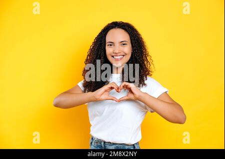 Bella candida piacevole latino o giovane donna brasiliana, in t-shirt di base, fa il gesto del cuore con le mani, dimostra segno d'amore, si alza su sfondo giallo isolato, guarda la macchina fotografica, sorrisi amichevole Foto Stock