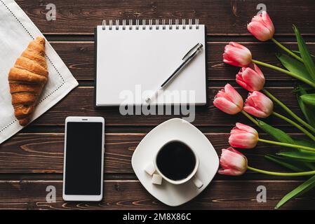 Tazza di caffè con un croissant per la prima colazione sul tavolo decorato con un bouquet di tulipani rosa e uno smartphone. Modello di biglietto d'auguri di primavera Foto Stock