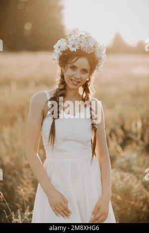 ritratto di sorridente bella ragazza in abito bianco con corona floreale e trecce in stile boho in estate al tramonto sul campo. Aggiunto un piccolo gr Foto Stock