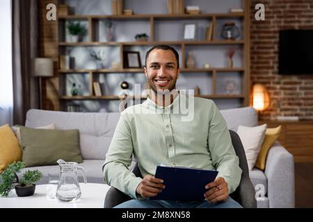 Giovane afro-americano seduto a casa in un elegante ufficio. Psicologo, allenatore o uomo d'affari che guarda la telecamera e sorridendo. Denti bellissimi Foto Stock