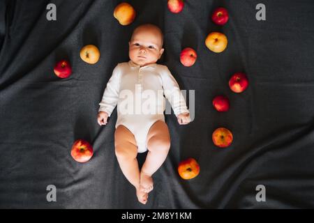 Neonato sdraiato con le mele. Un bambino piccolo in un bodysuit bianco su uno sfondo nero. Vista dall'alto, disposizione piatta Foto Stock