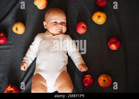 Neonato sdraiato con le mele. Un bambino piccolo in un bodysuit bianco su uno sfondo nero. Vista dall'alto, disposizione piatta Foto Stock