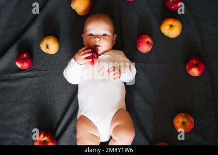Neonato sdraiato con le mele. Un bambino piccolo in un bodysuit bianco su uno sfondo nero. Vista dall'alto, disposizione piatta Foto Stock