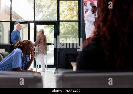 Fuoco selettivo di un assistente medico esausto stanco seduto sulla sedia nell'area di attesa dell'ospedale che ha una pausa prima di iniziare l'esame. Infermiera afroamericana che lavora al trattamento di salute del paziente Foto Stock