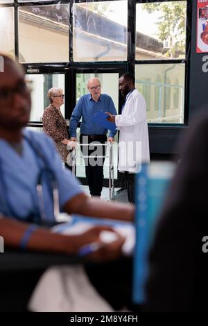 Medico curante che discute i sintomi della malattia con paziente anziano ferito prima di iniziare la consultazione medica. Vecchio con telaio per camminare che ha appuntamento in ospedale sala d'attesa Foto Stock