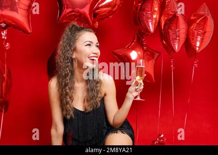 Felice giovane donna con rossetto rosso che tiene in vetro champagne seduta su sfondo rosso in abito nero con palloncini ragazza caucasica celebra il compleanno di festa o San Valentino con ampio sorriso toothy Foto Stock