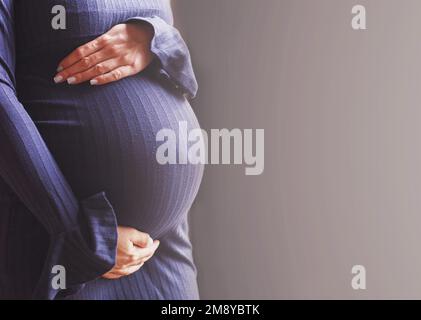 il primo piano della pancia di una donna incinta. Le mani delle ragazze abbracciano il ventre su sfondo blu. Concetto di salute materna. Foto Stock