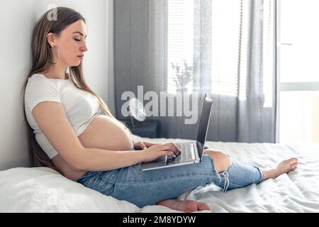 Attraente donna incinta si trova sul letto e utilizza un computer portatile. Foto Stock