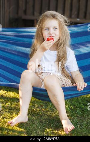 la bambina bionda con i capelli lunghi mangia una fragola nel giardino seduta in un'amaca. Foto Stock