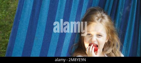 la bambina bionda con i capelli lunghi mangia una fragola nel giardino sdraiato in un'amaca. Foto Stock