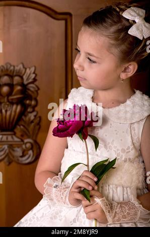 Vacanza per bambini. Ragazza elegante in un abito bianco. Ragazza con bouquet di peonie fresche su sfondo di legno Foto Stock