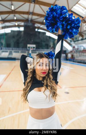 Scatto medio. Una cheerleader bionda in uniforme, con un braccio dietro la testa e l'altro allungato. Concetto sportivo. Foto verticale. Foto di alta qualità Foto Stock